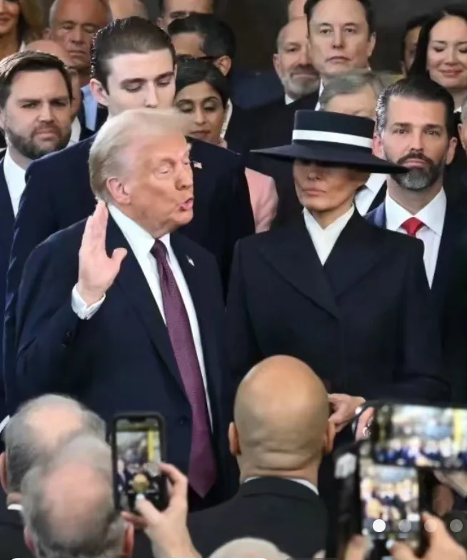A photo of Donald Trump speaking at a public event, gesturing with his right hand, surrounded by a crowd including Elon Musk…