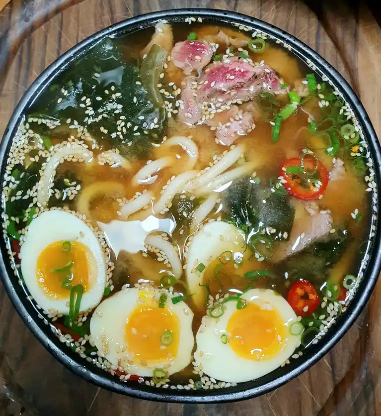 A top-down photo of a bowl of ramen with udon noodles, sliced meat, seaweed, green onions, sesame seeds, and four halves of…