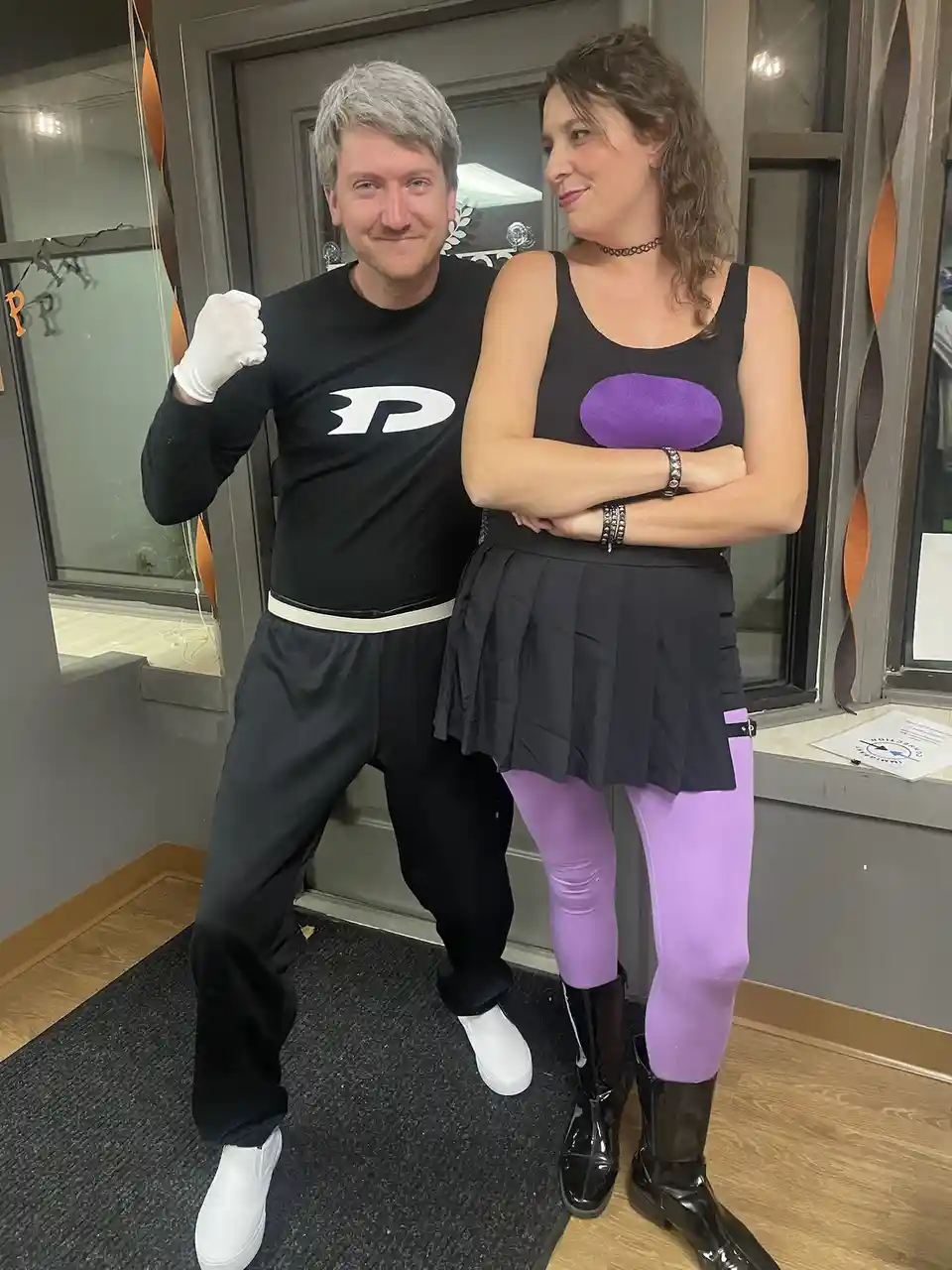 A man and a woman pose together indoors. The man, on the left, wears a black long-sleeved shirt with a white 'P' logo, black…