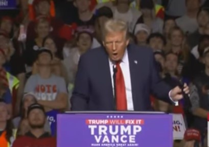 A man in a dark suit and red tie stands at a purple podium with 'TRUMP VANCE' and 'MAKE AMERICA GREAT AGAIN!' text. Behind…