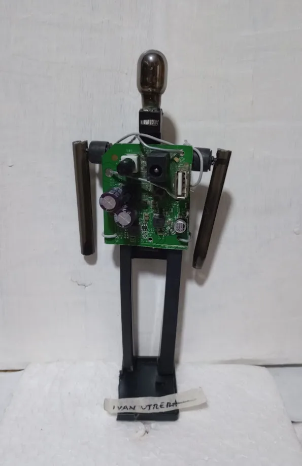 A handmade robot figure constructed from a green circuit board for a torso, with a glass bulb for a head, metal rods for…