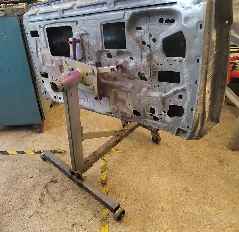 A large, bare metal car body panel, likely a door or fender, is mounted on a wheeled metal stand in a workshop. The panel…
