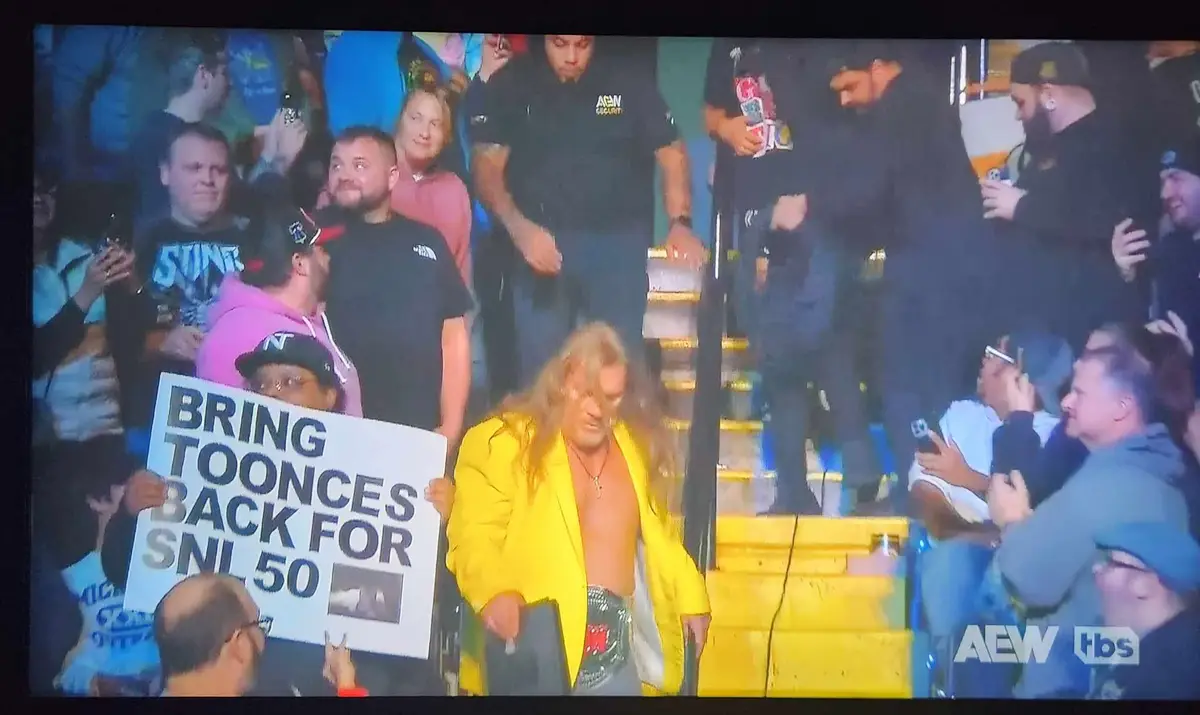 A man with long blonde hair and a yellow jacket walks down yellow steps surrounded by a crowd. One person holds a sign that…