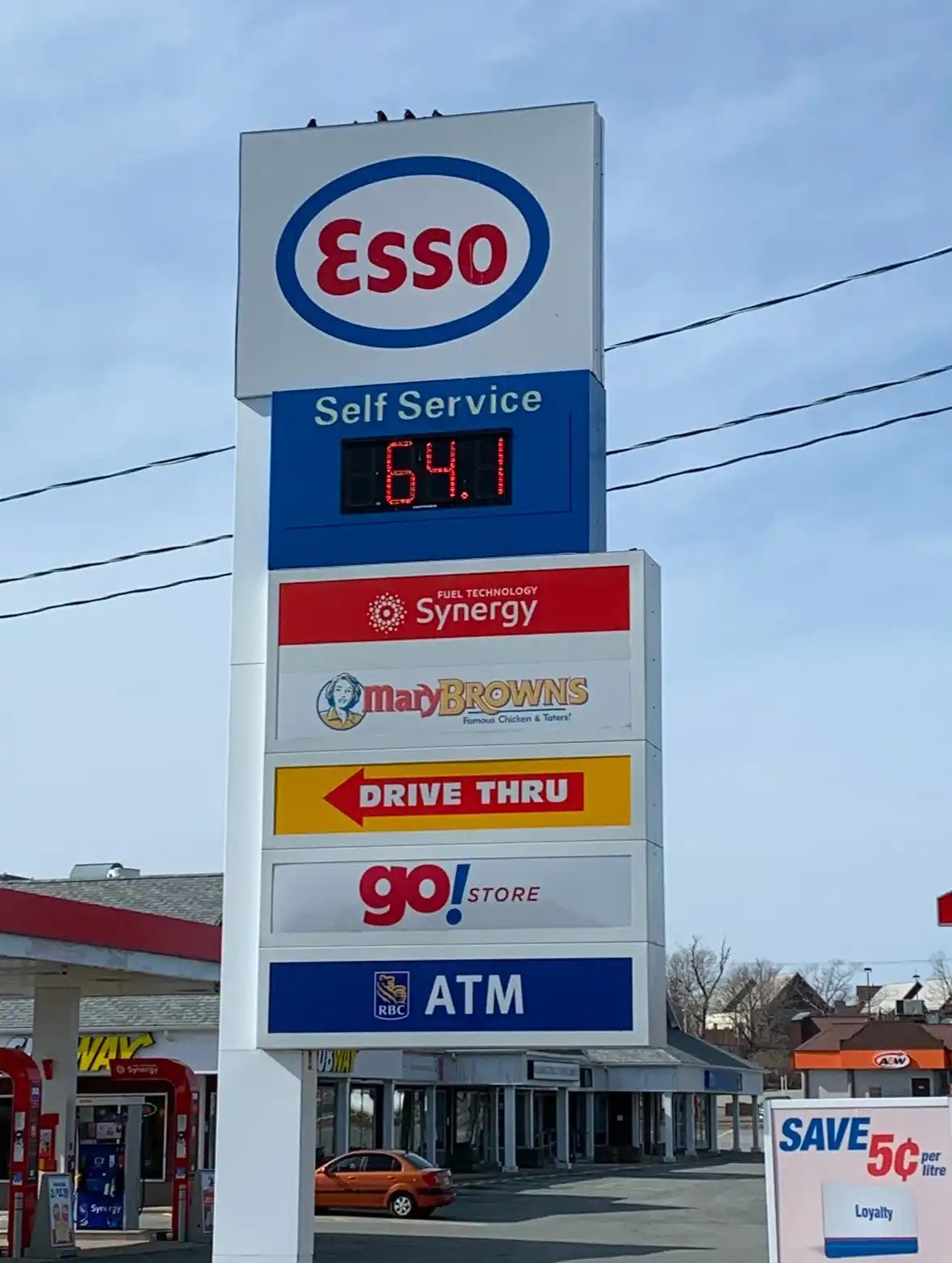 A tall Esso gas station sign displays a digital price of 64.1 cents per litre under 'Self Service', with logos for Synergy…