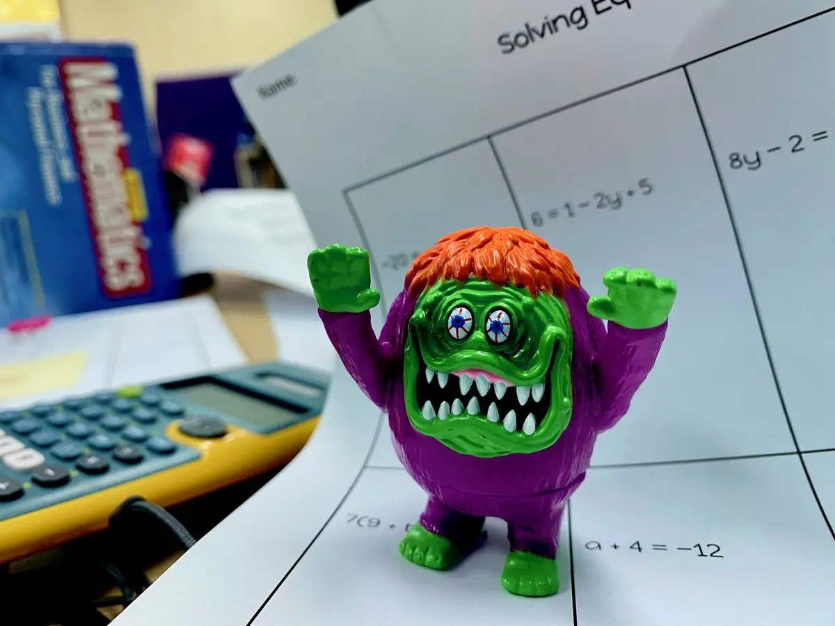 A green, purple, orange-haired monster toy with wide eyes and raised fists stands triumphantly on a math worksheet filled…
