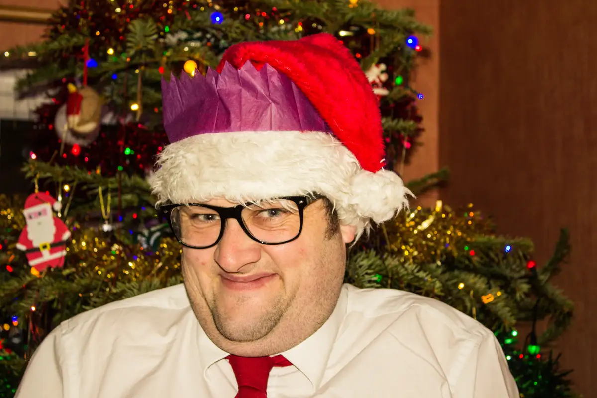 A man wearing a red Santa hat with a purple paper crown on top, black-rimmed glasses, a white collared shirt, and a red tie…