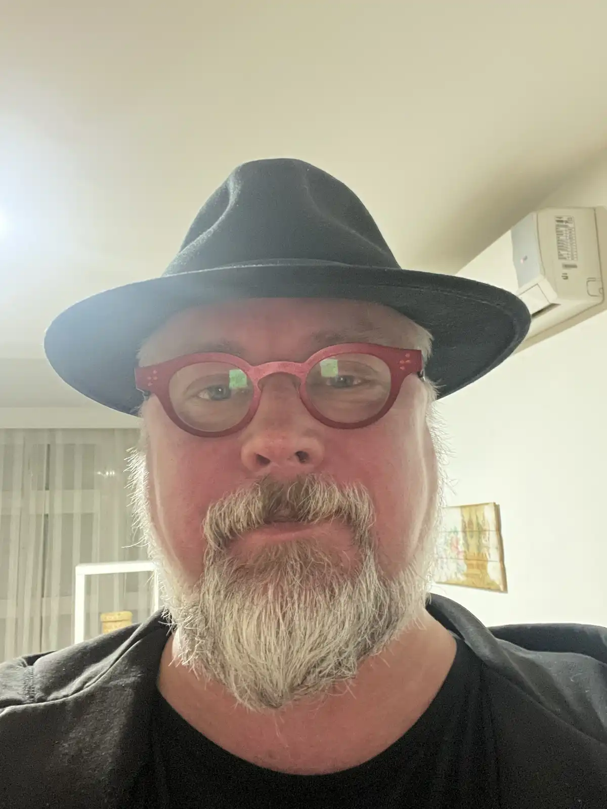 A close-up selfie of a middle-aged man with a gray beard and mustache, wearing a black fedora and bright red-rimmed glasses…