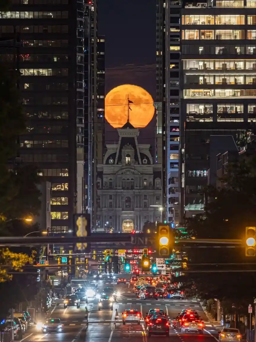A nighttime city street view framed by tall buildings, with a large, orange, full moon appearing to sit directly behind a…