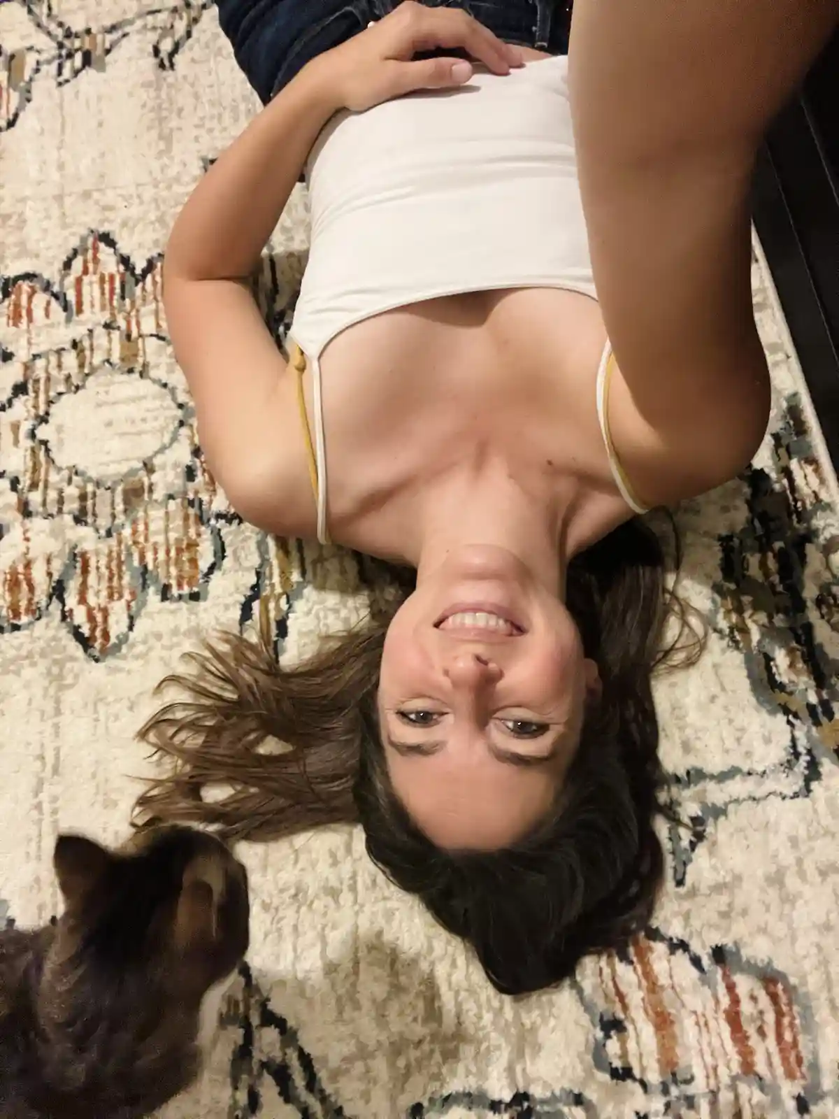A woman lies on her back on a patterned rug, smiling up at the camera while wearing a white tank top and jeans. A…