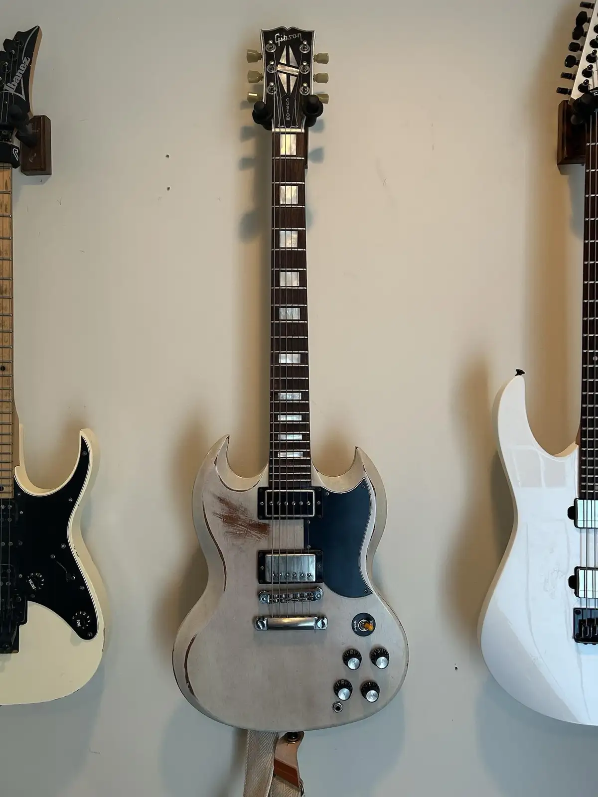Three electric guitars hang on a wall, with a central Gibson Custom SG in a worn, light-colored finish. The text overlaid on…