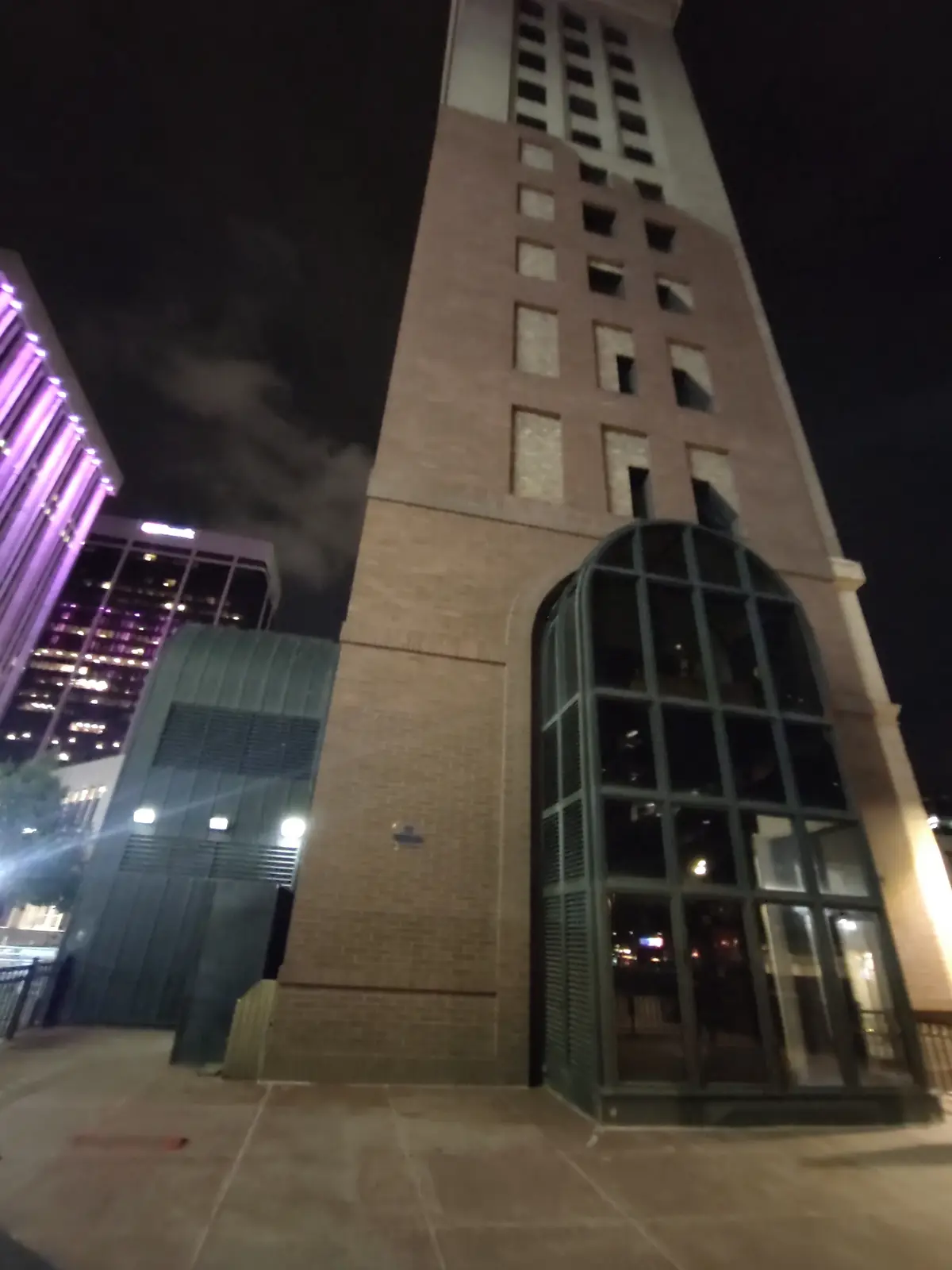 A low-angle nighttime photo shows a tall brick building with an arched glass entrance, next to a modern building with purple…