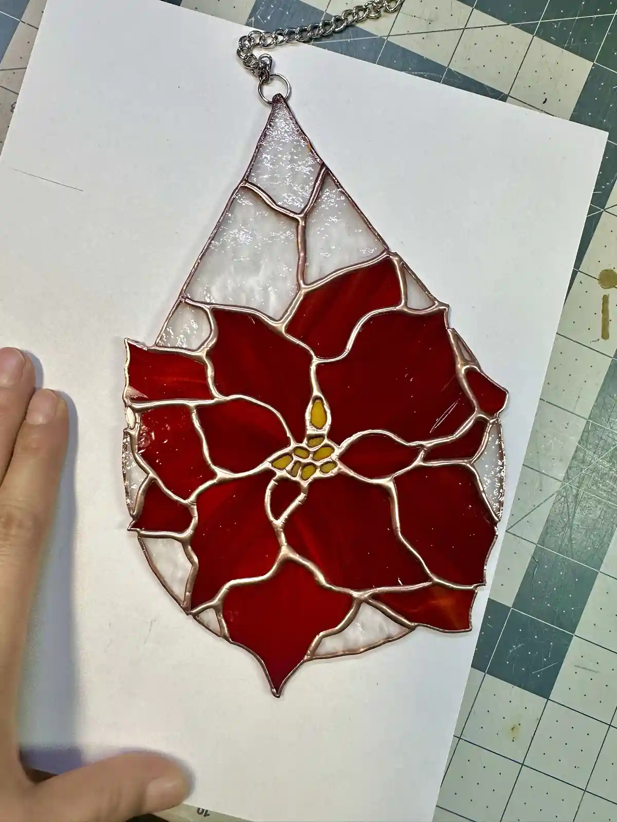 A hand holds a white sheet of paper displaying a stained glass pendant shaped like a poinsettia flower, featuring red…