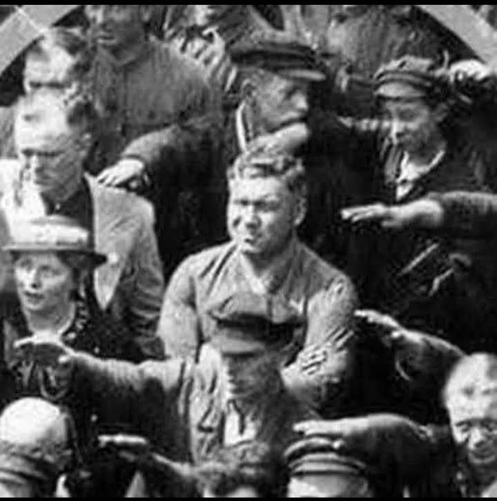 A grainy black-and-white historical photograph shows a man in the center of a crowd, surrounded by other people, with his…