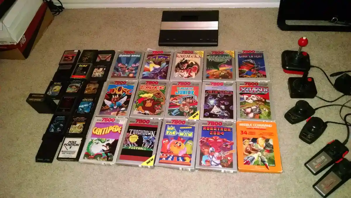 An Atari 7800 video game console sits on a carpeted floor surrounded by dozens of its game cartridges, including titles like…
