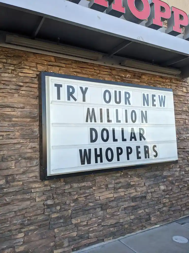 A white marquee sign mounted on a stone facade of a restaurant, with black letter tiles spelling 'TRY OUR NEW MILLION DOLLAR…
