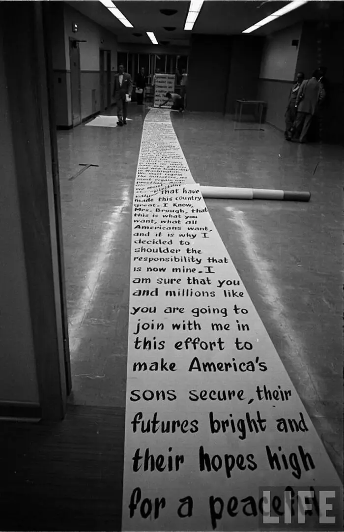 A black-and-white photo shows a long strip of paper with handwritten text laid out on a hallway floor, leading toward the…