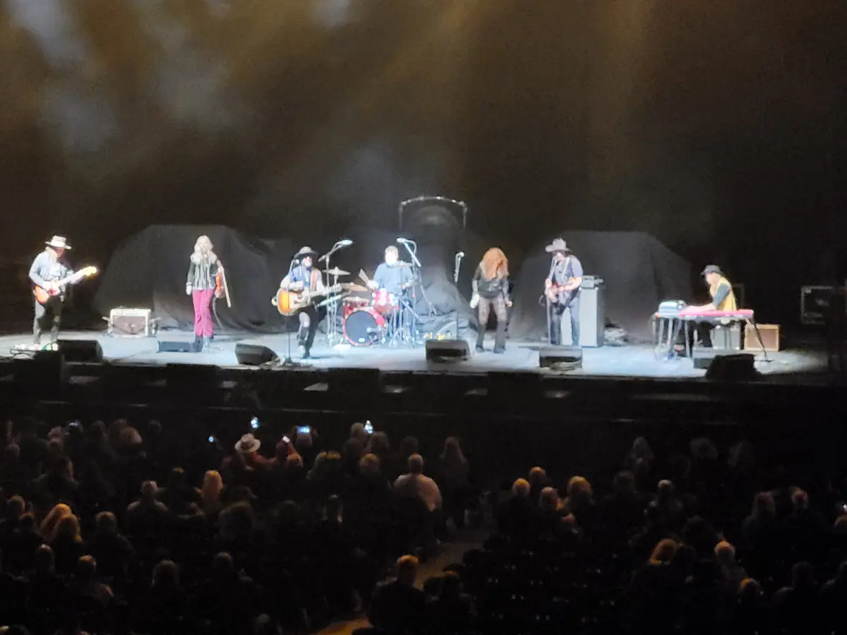 A live band performs on a dimly lit stage with a large audience in the foreground. Seven musicians are visible, including…