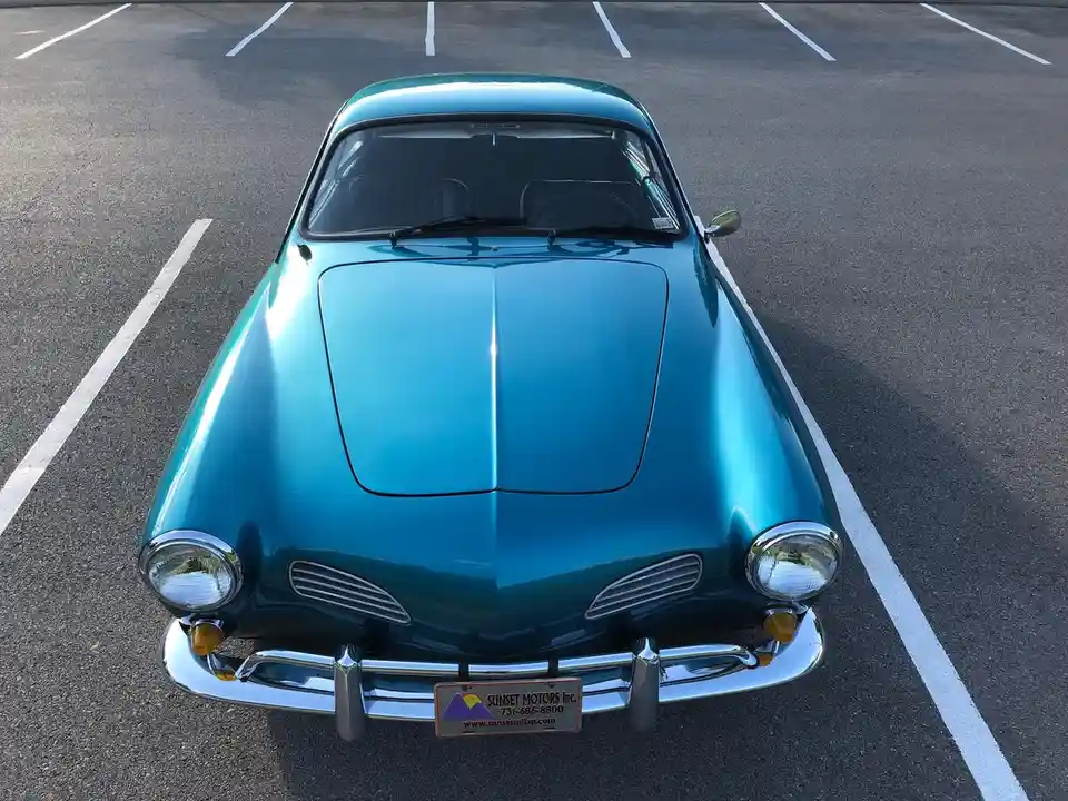 A high-angle front view of a shiny teal vintage car parked in an asphalt lot marked with white parking lines. The car has a…