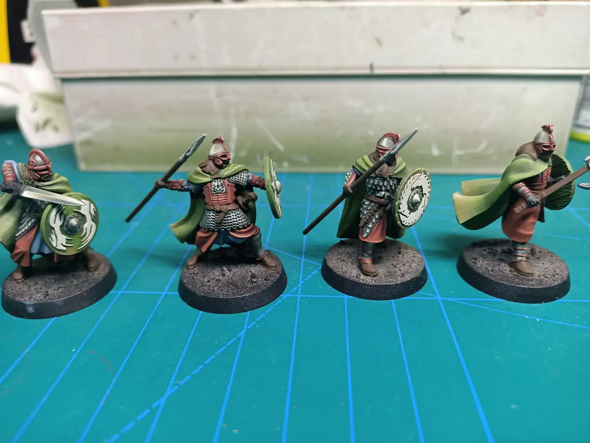 Four hand-painted miniature fantasy warriors in armor and green cloaks stand on circular bases on a blue grid mat. Each…