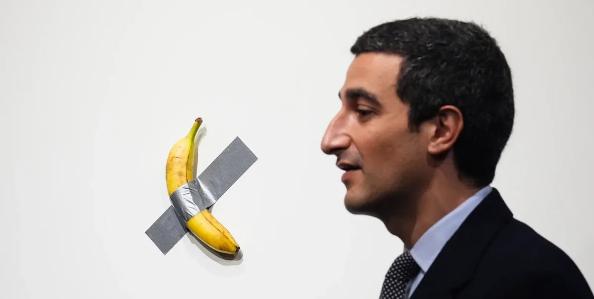 A man in a suit looks at a banana taped to a white wall with duct tape in an X shape. The banana is yellow with some brown…