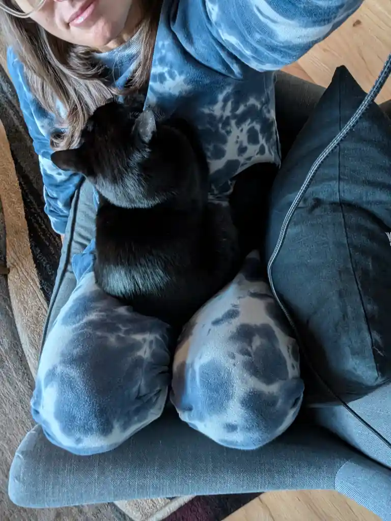 A person wearing a blue tie-dye hoodie and matching pants sits on a gray couch, smiling slightly while holding a black cat…