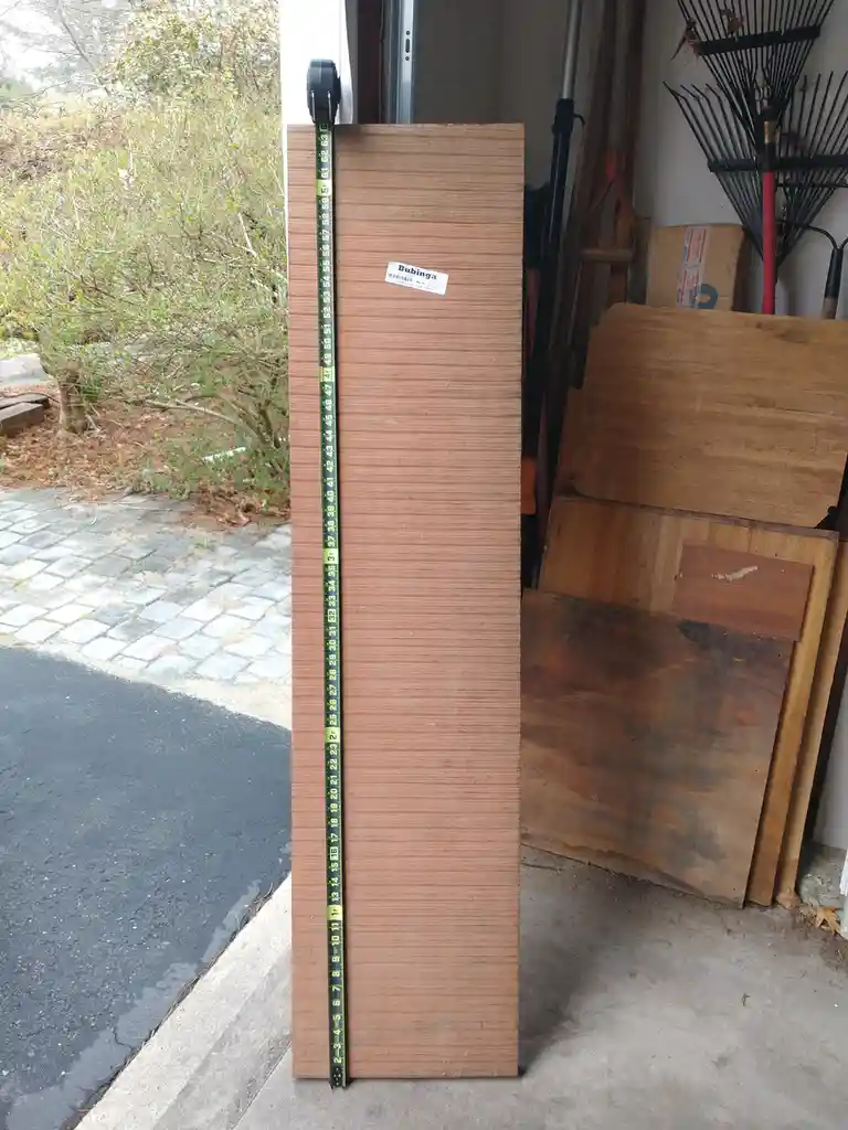 A tall, reddish-brown wooden board labeled 'Bubinga' stands vertically in a garage, with a yellow measuring tape stretched…