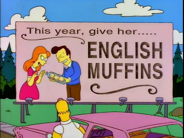 A billboard from The Simpsons shows a man in a blue sweater giving a tray of English muffins to a woman in a pink dress…