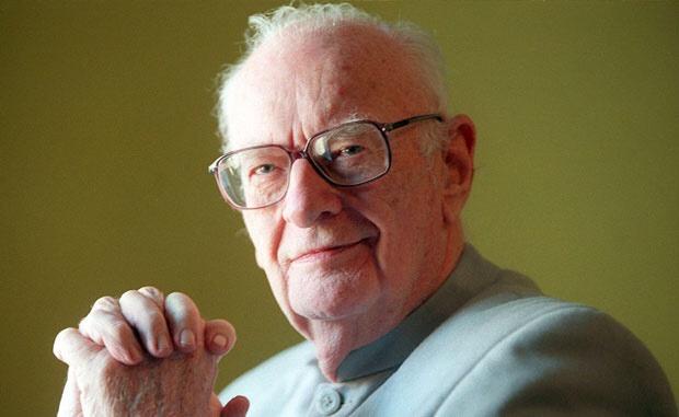 An elderly man with white hair and glasses, wearing a light-colored collared shirt, looks directly at the camera with a…
