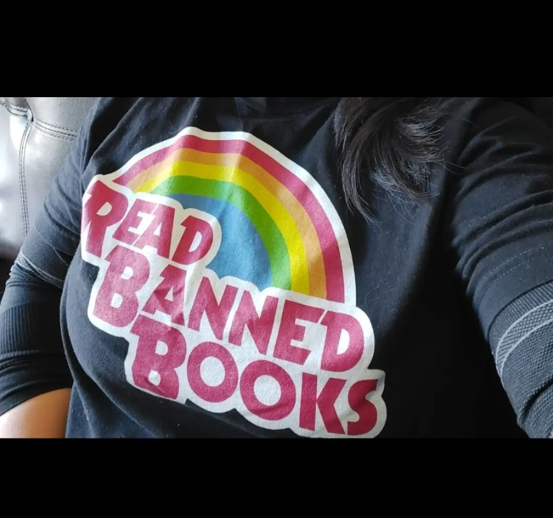 A close-up photo shows a person wearing a black long-sleeved shirt with a colorful rainbow graphic and bold pink text that…