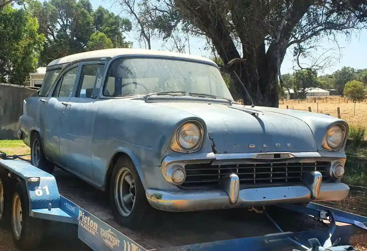 A weathered, light blue vintage Holden sedan, showing signs of rust and age, is parked on a blue flatbed trailer labeled…
