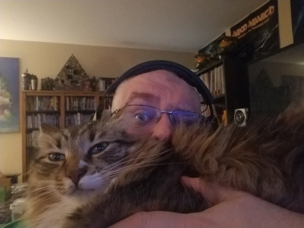 A man wearing glasses and headphones holds a fluffy, long-haired tabby cat close to his face in a selfie. The cat looks…