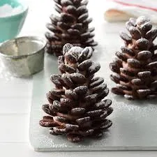 Three chocolate-covered pinecone-shaped cookies dusted with powdered sugar sit on a white cutting board, with a small metal…