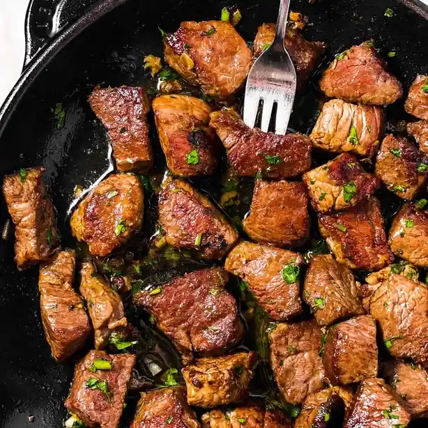 a close-up photo of diced steak pieces sizzling in a black cast iron skillet, garnished with chopped green herbs and a…