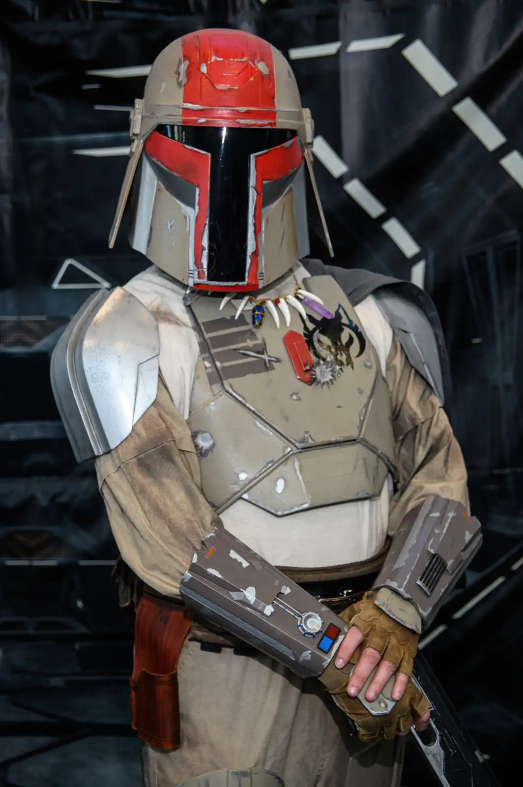 A person in a detailed, weathered Mandalorian armor costume with a red and tan helmet, holding a blaster. The armor has…