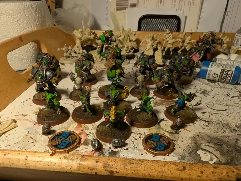 A cluttered, paint-splattered workbench displays a collection of painted green orc miniatures with armor and weapons…