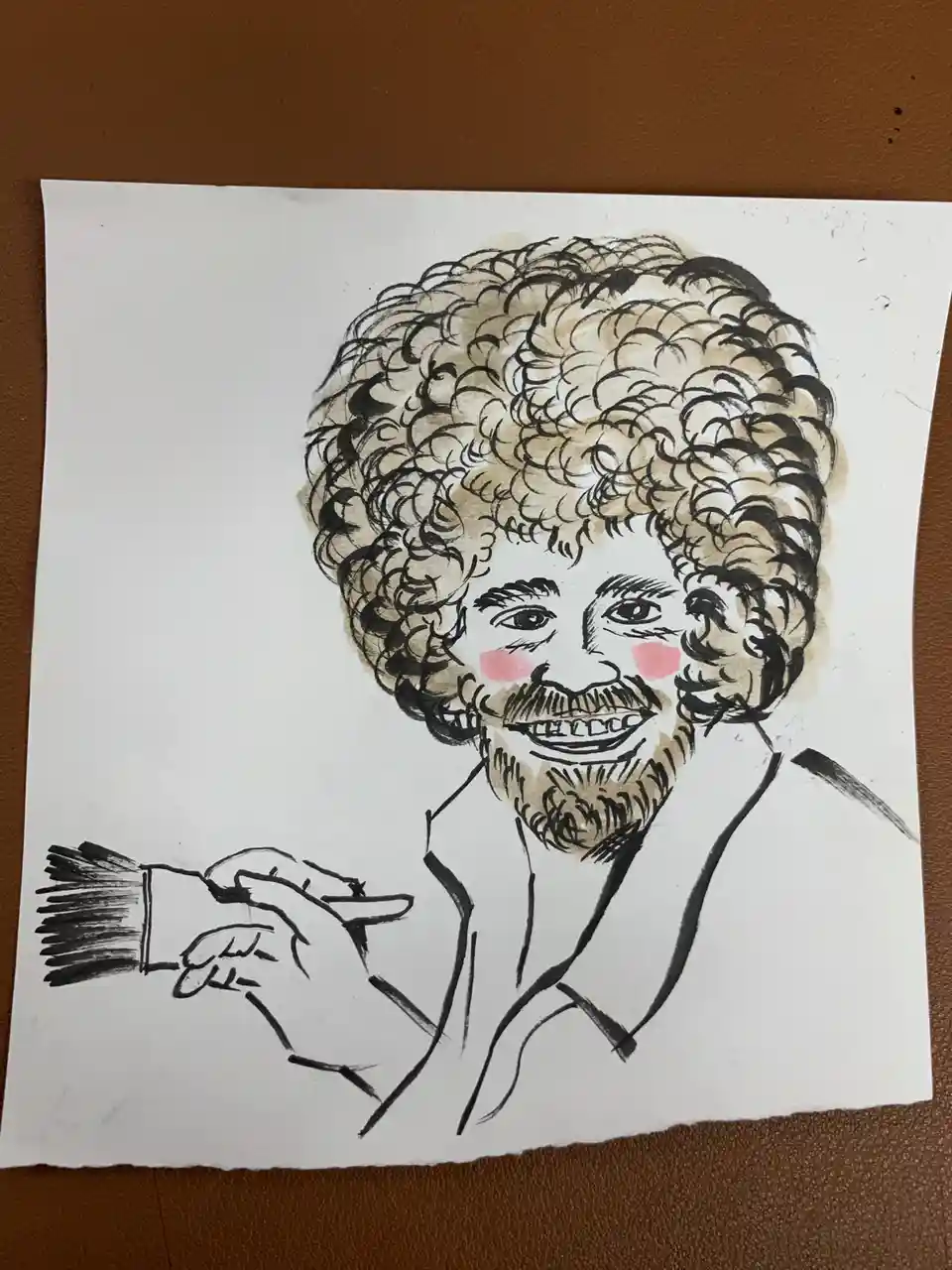A hand-drawn portrait on paper depicts a smiling man with a large, curly afro, rosy cheeks, and a beard, holding a…
