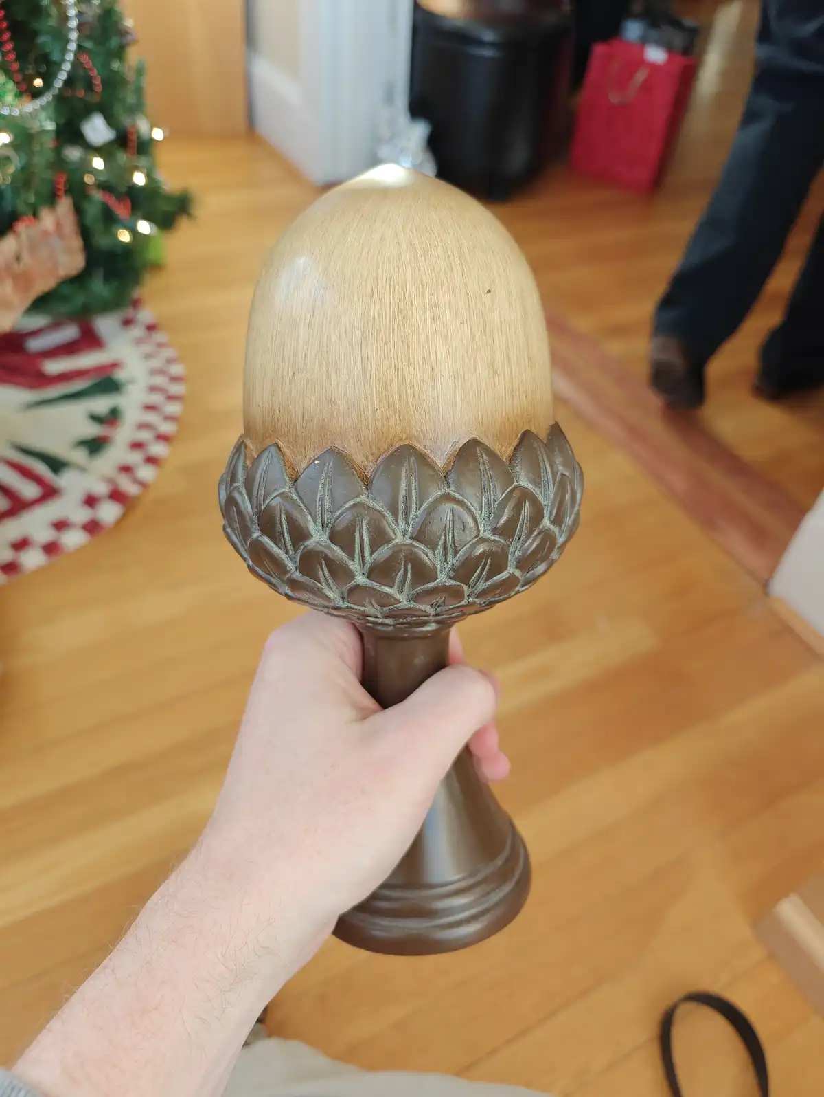 A hand holds a decorative, acorn-shaped object that appears to be a Christmas tree stand, with a light wood top and dark…