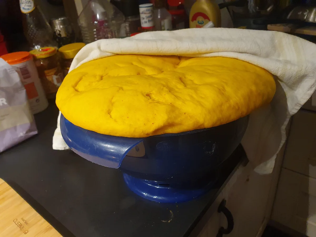 A large, vibrant yellow dough has risen dramatically to overflow the rim of a blue plastic mixing bowl, covered partially by…