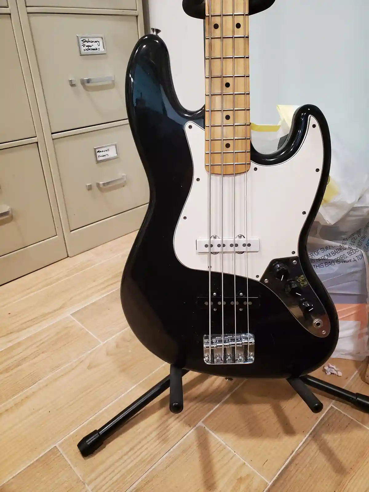 A black and white electric bass guitar with four strings stands upright on a black tripod stand on a light wood floor. In…