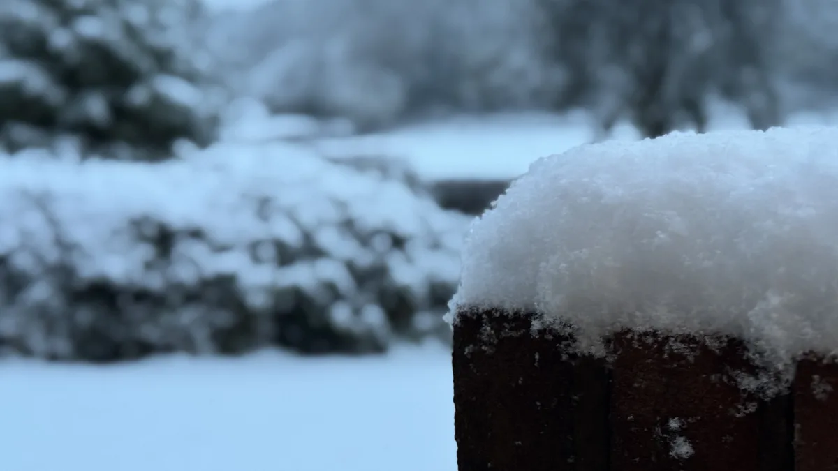 A close-up photo shows a thick, fluffy layer of snow resting on the top of a dark brown wooden fence post. The background is…