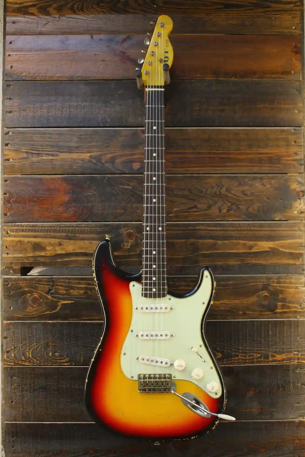 A sunburst-finish electric guitar with a light green pickguard is mounted vertically against a dark wooden plank wall. The…
