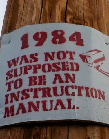 A white sign with red text is taped to a wooden utility pole. The text reads '1984 WAS NOT SUPPOSED TO BE AN INSTRUCTION…