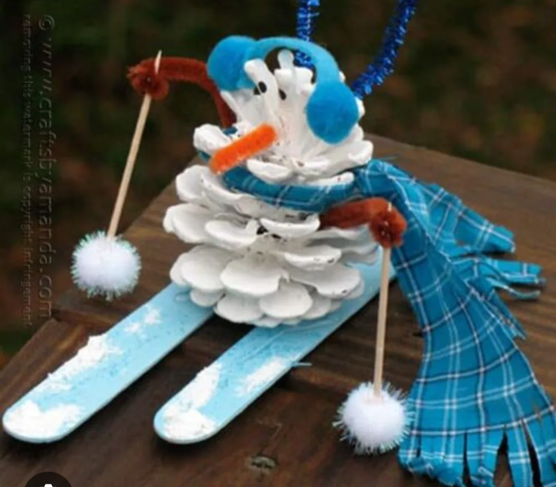 A handmade pinecone snowman craft sits on blue skis, wearing blue earmuffs, a blue plaid scarf, and holding two ski poles…