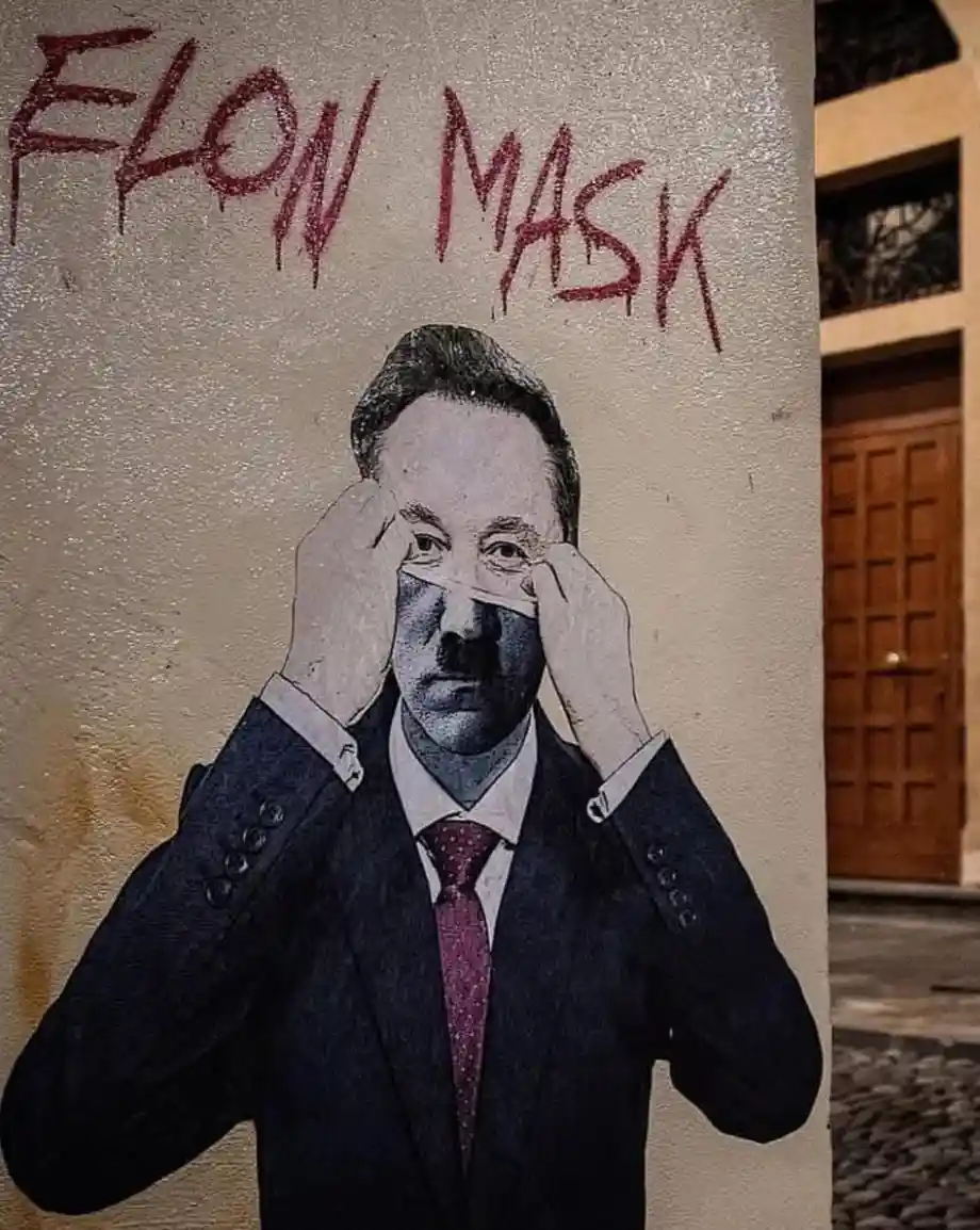 A street mural depicts a man in a suit and tie, holding a white strip across his eyes like a mask. Above the figure, red…