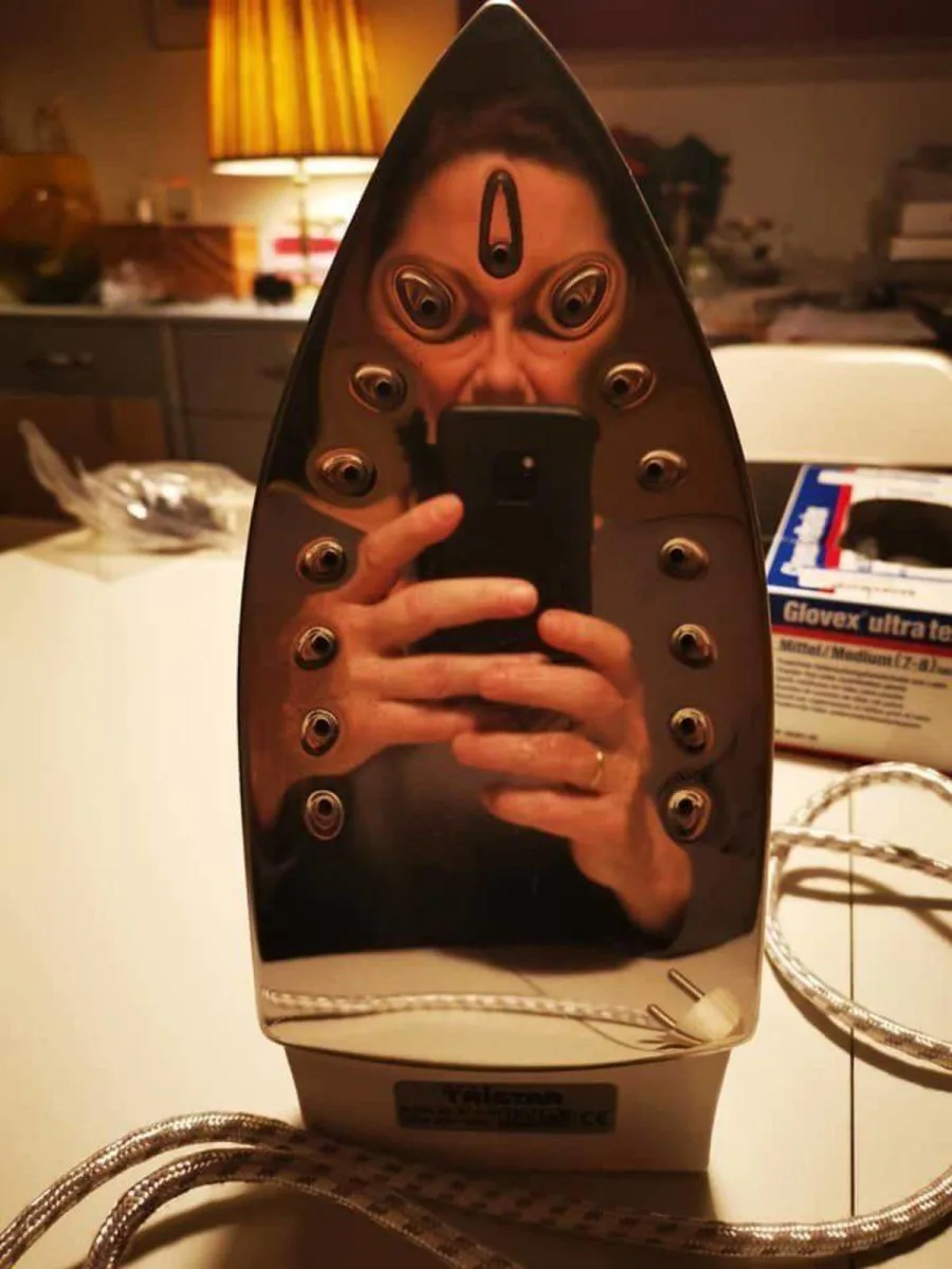 A shiny, metallic steam iron sits upright on a countertop, reflecting a distorted, unsettling image of a person taking a…