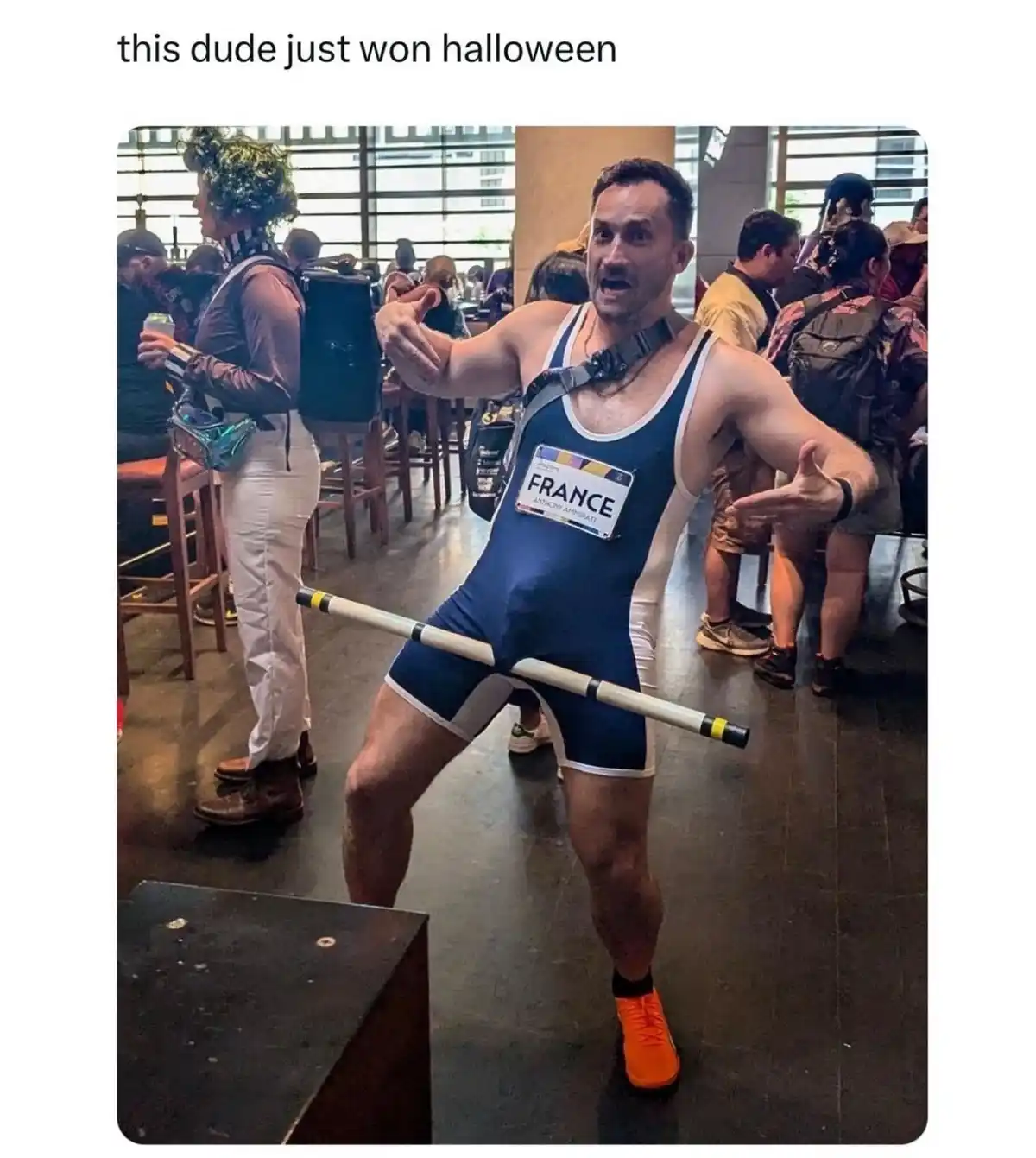 A man in a blue and white wrestling singlet with a 'FRANCE' name tag stands triumphantly in a crowded indoor space, holding…