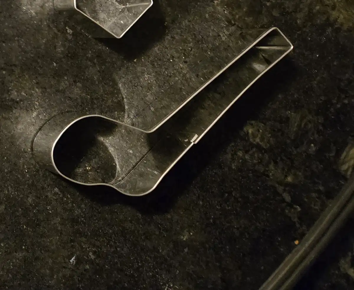 A metal cookie cutter shaped like a spoon lies on a dark, speckled countertop, with another cookie cutter partially visible…