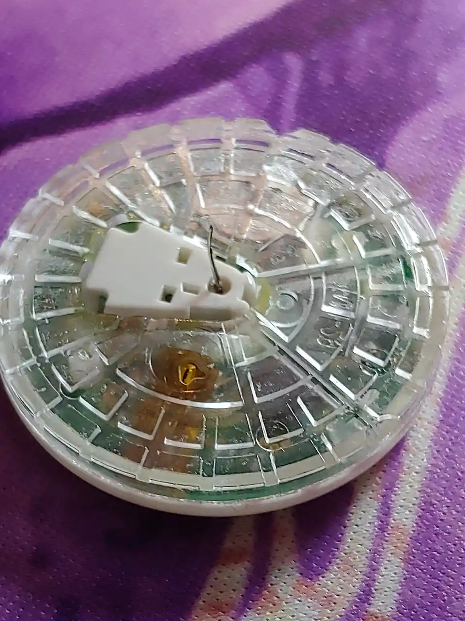 A broken watch with its glass face shattered into a mosaic pattern, revealing the internal mechanism and a white plastic…