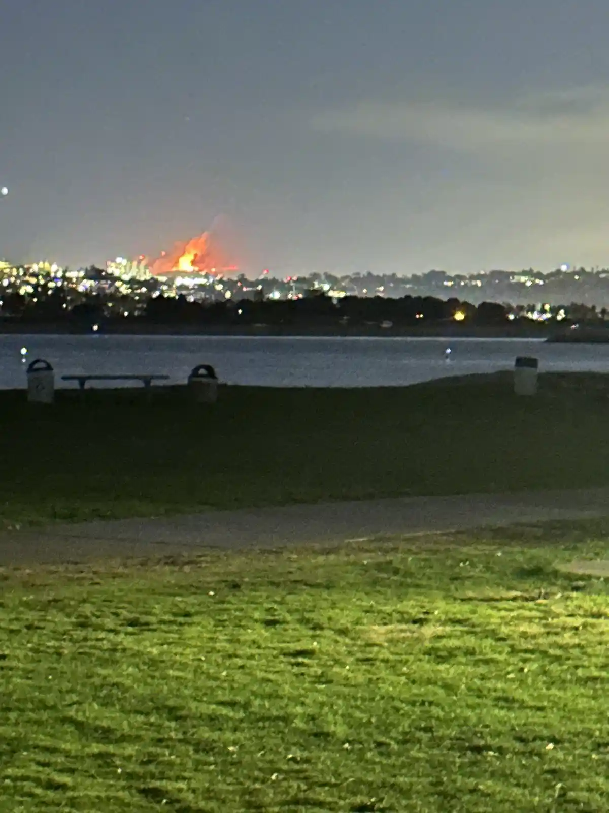 A nighttime photo from a grassy park overlooking water, with a distant cityscape and a large fire burning on the horizon…