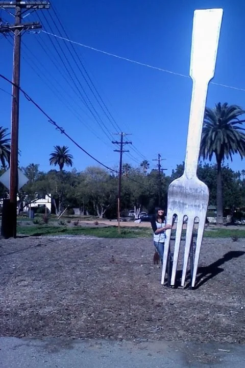A person stands beside a giant white plastic fork sculpture outdoors under a clear blue sky, with palm trees and utility…